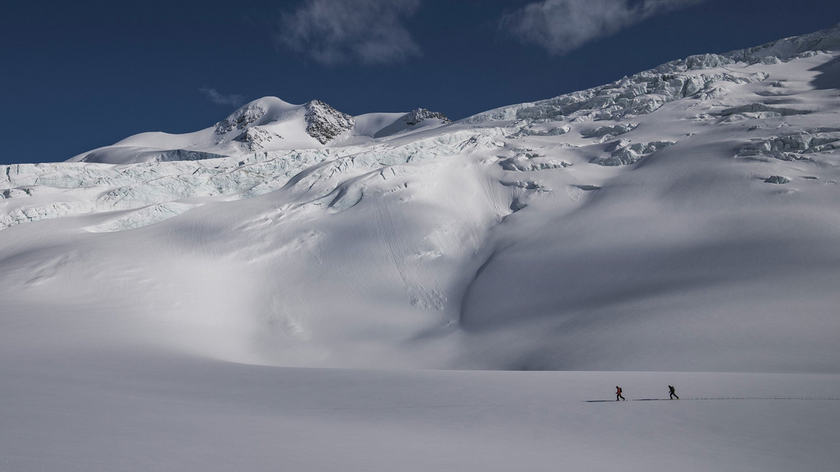 A wide snowy mountain landscape under a deep blue sky, with two tiny distant figures on the snow in the lower right.