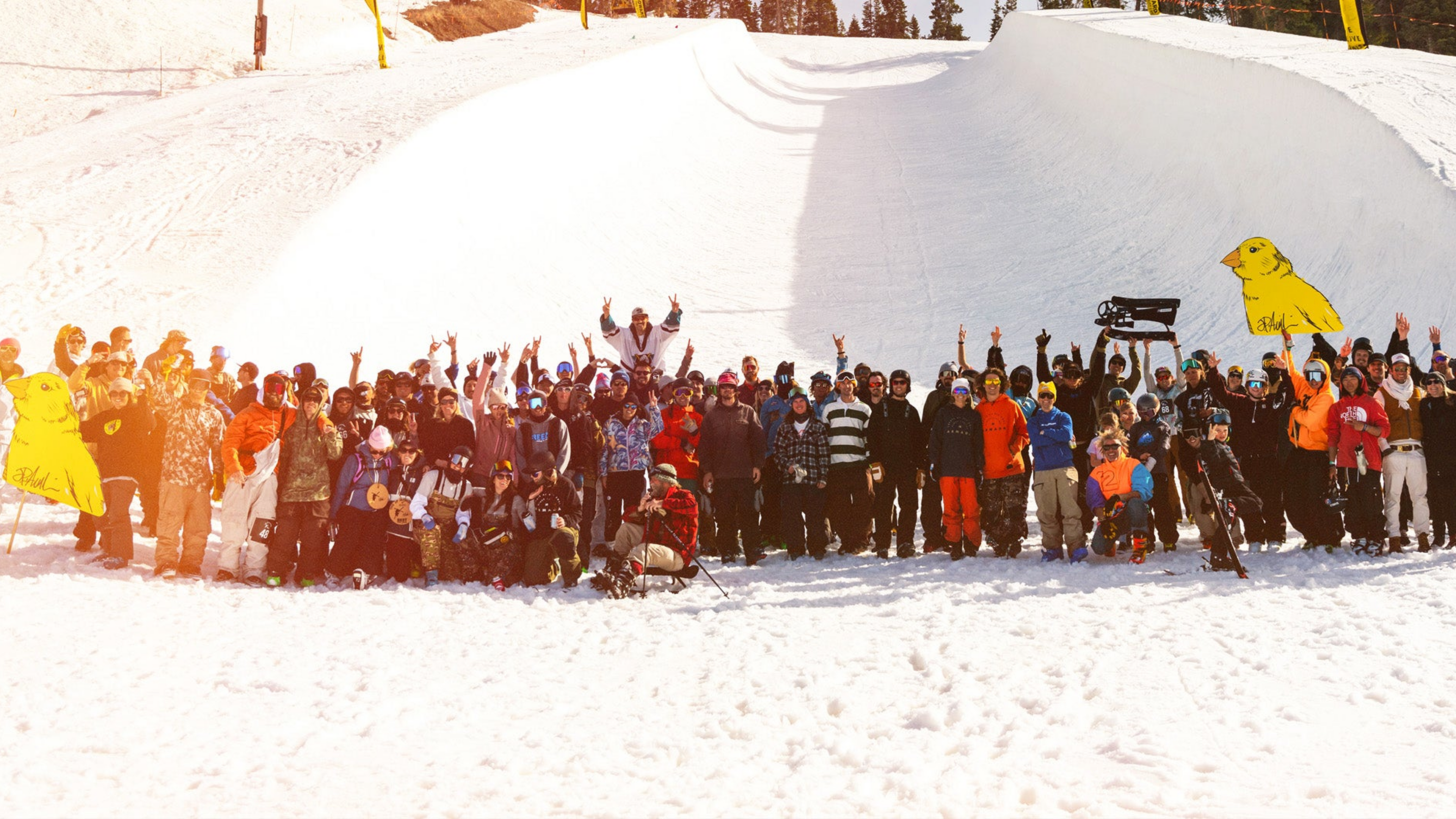 A large group of snowboarders and skiers posing together on a snowy mountain slope with a large ramp in the background. The group is dressed in various colorful winter sports gear, and a bright yellow bird-shaped sign is visible on the right side of the slope.