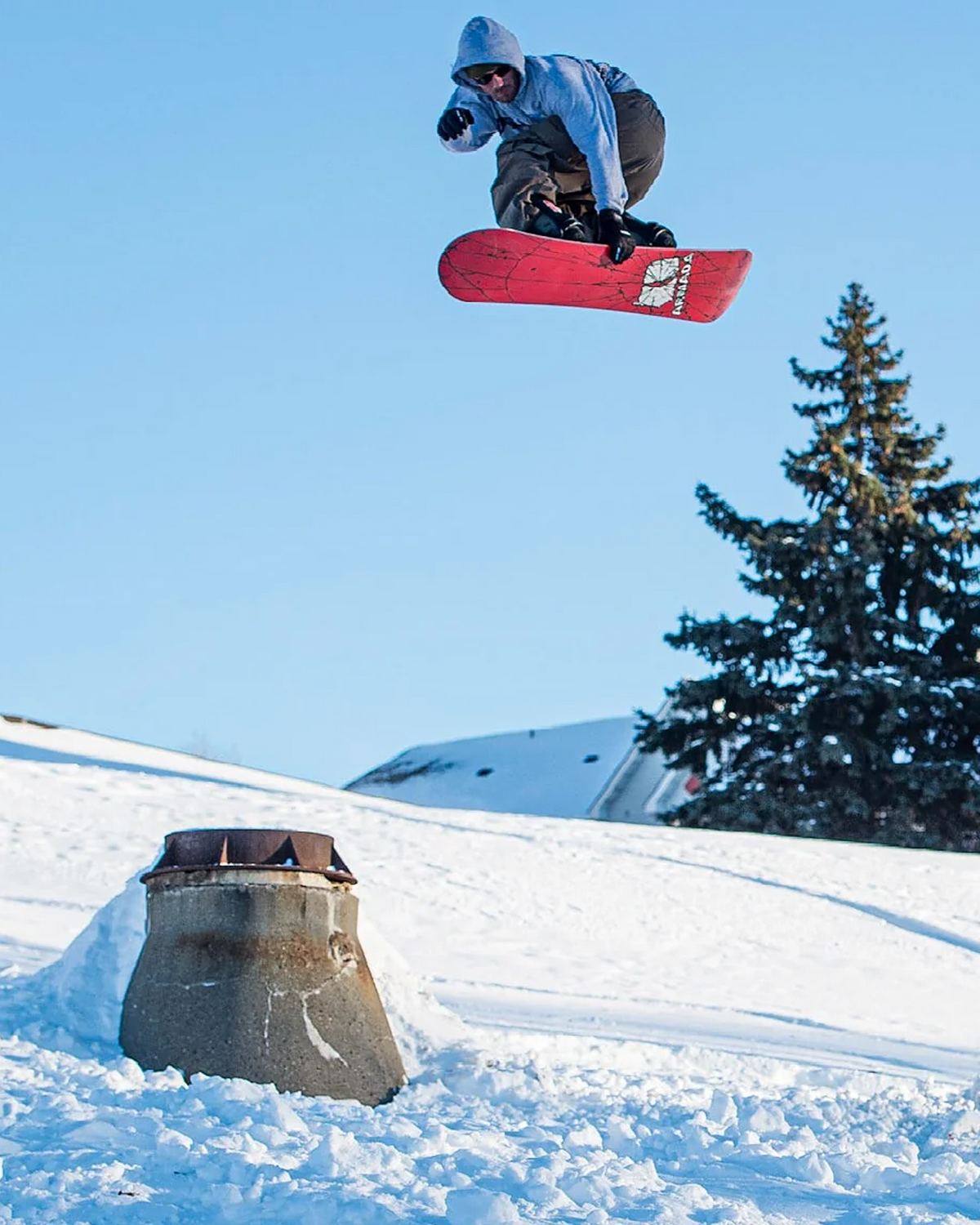 A snowboarder in a light blue jacket and dark pants jumps in the air over a snowy slope, holding a red snowboard with a visible white and black logo. A concrete jump ramp sits in the snow below, with a pine tree and blue sky in the background.