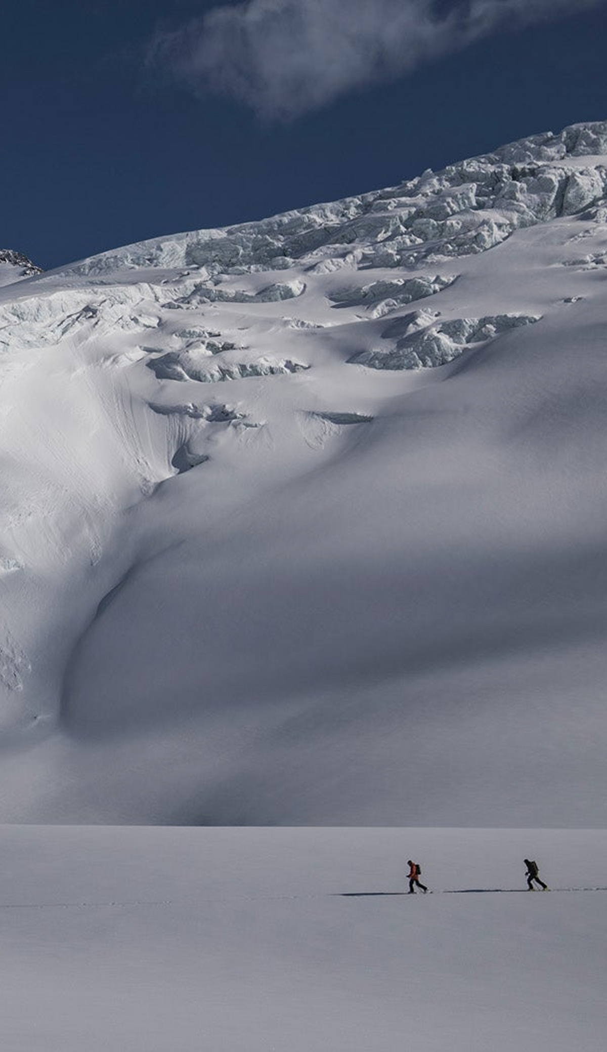 A vast snowy mountain landscape under a deep blue sky, with two tiny hikers visible near the bottom of the slope.