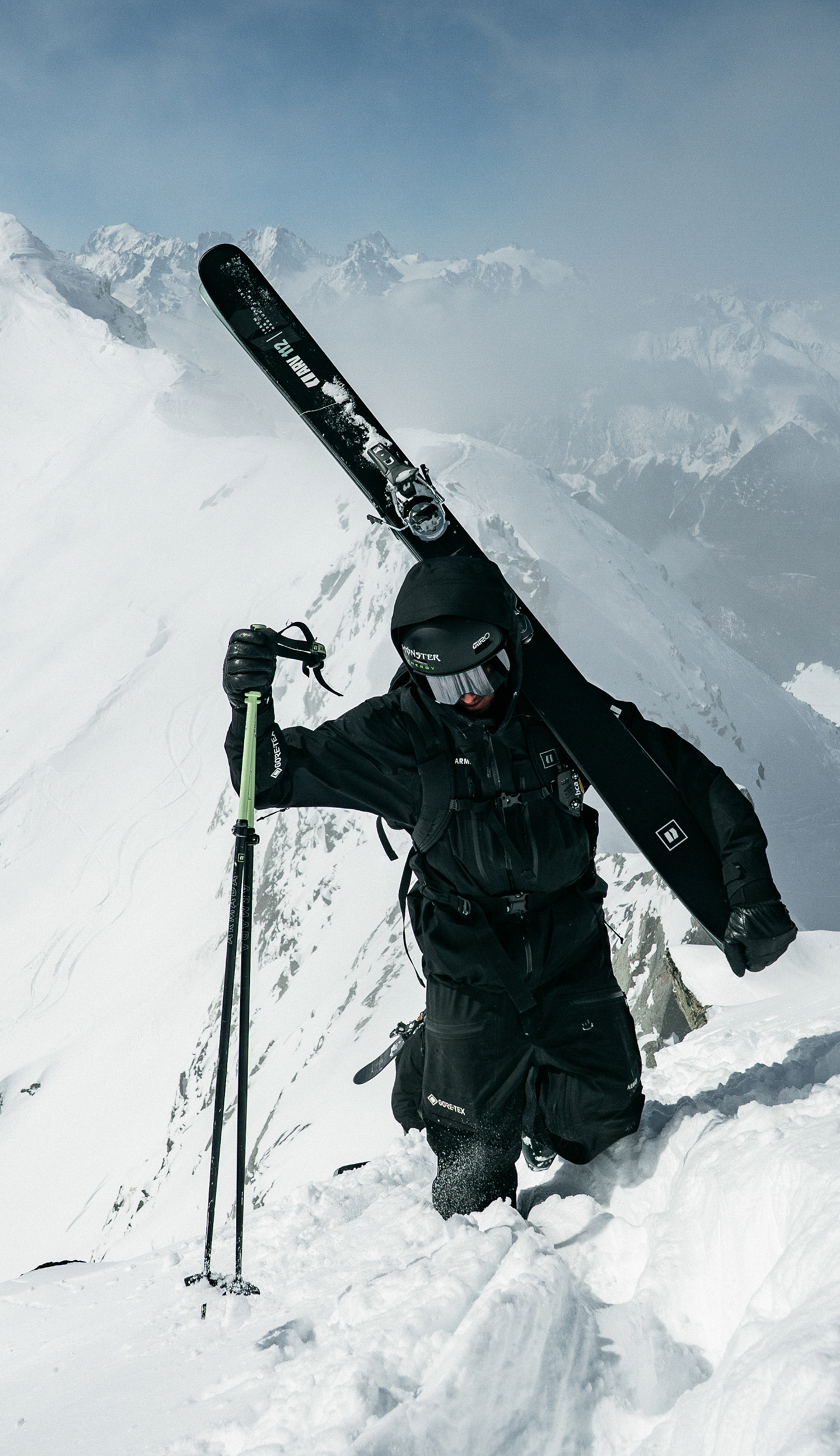 A skier in a black ski suit and helmet carries a pair of black skis on a snowy mountain slope, holding ski poles with mountains in the background.