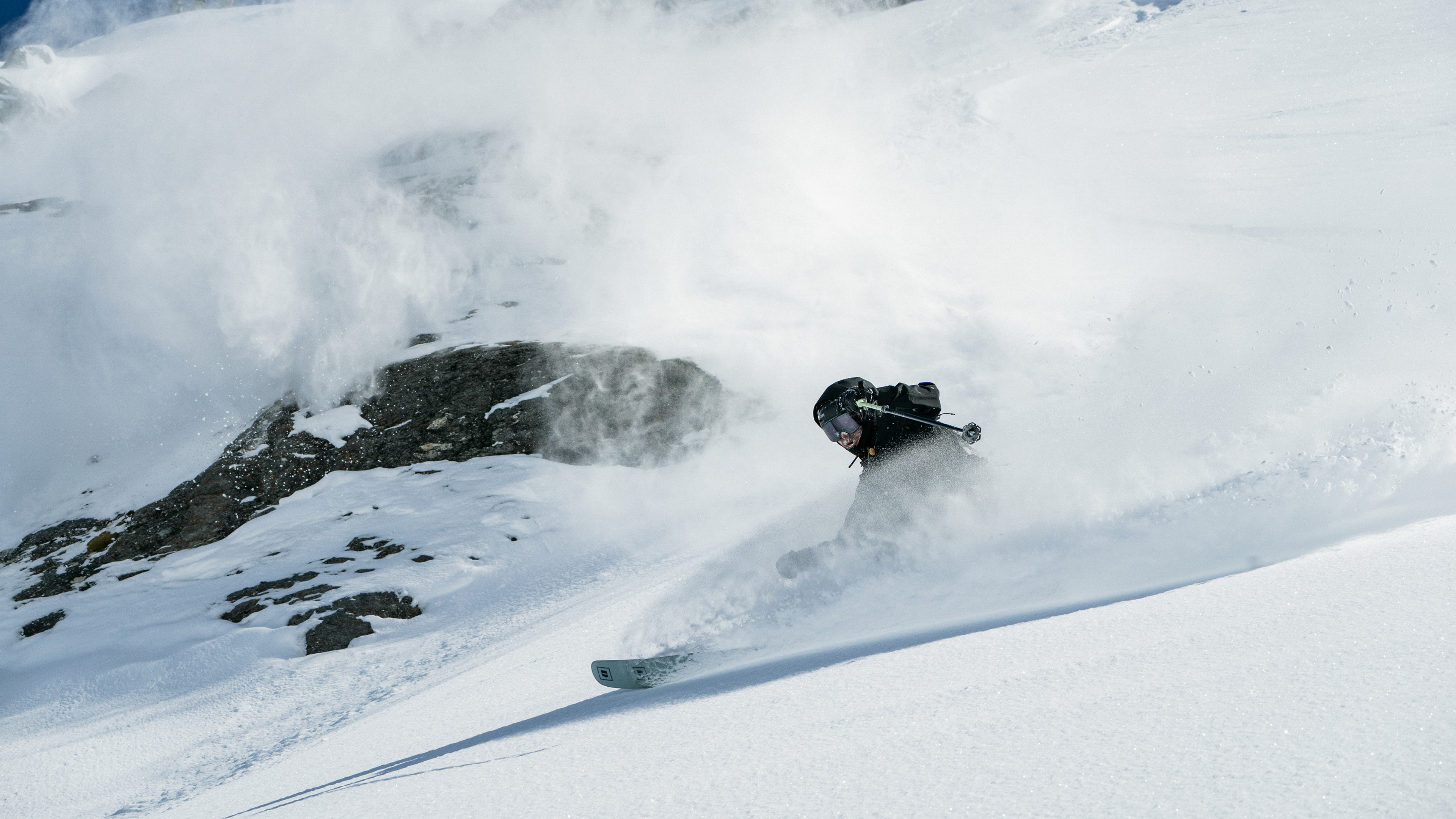 A skier carving through deep powder snow, kicking up a large spray of white snow and dusting dark rocks on a steep snowy slope.
