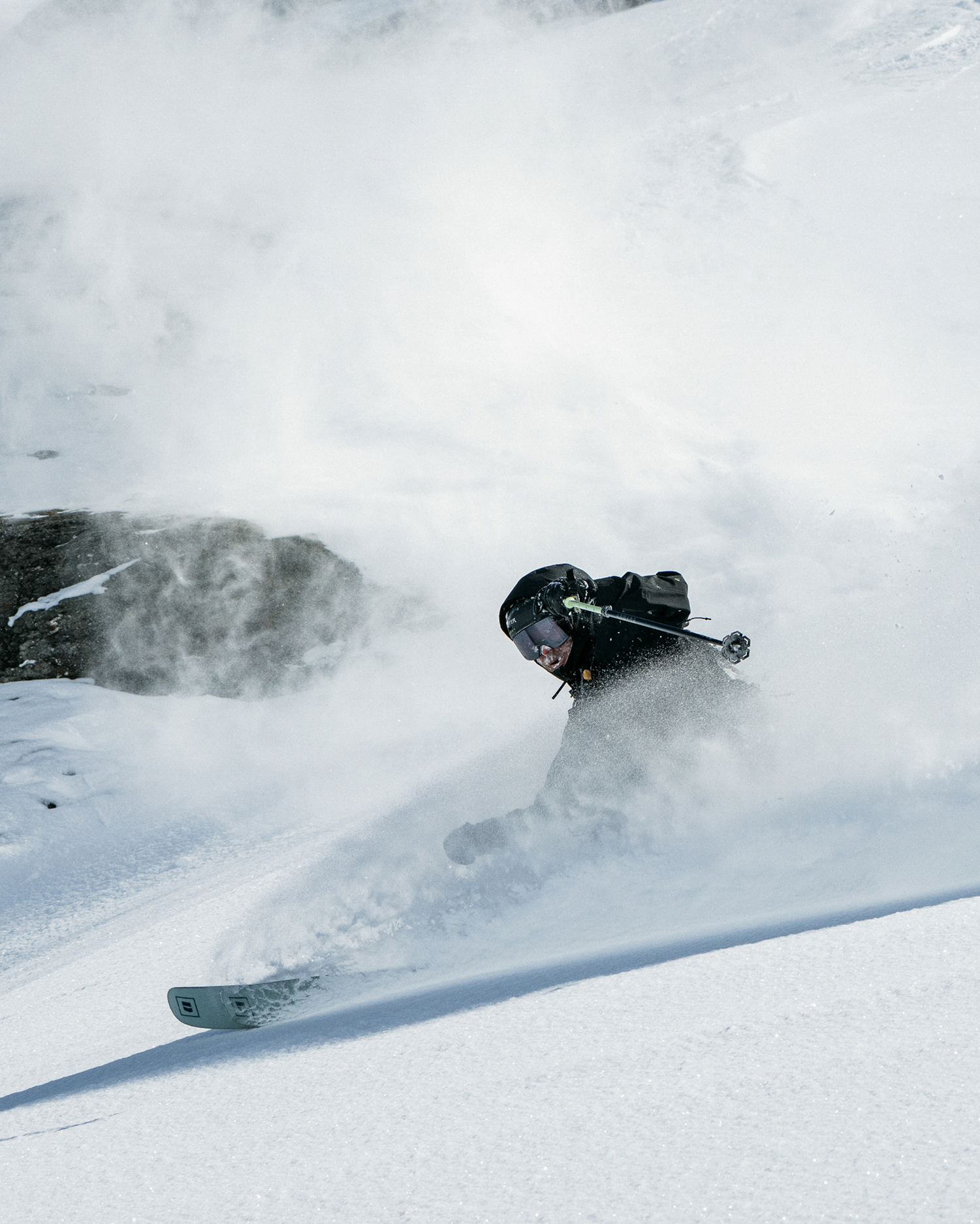 A skier carving through deep powder snow, kicking up a large spray of white snow. The skier is wearing a black jacket, black helmet, and goggles, with ski poles in one hand and a snowboard visible under their feet.