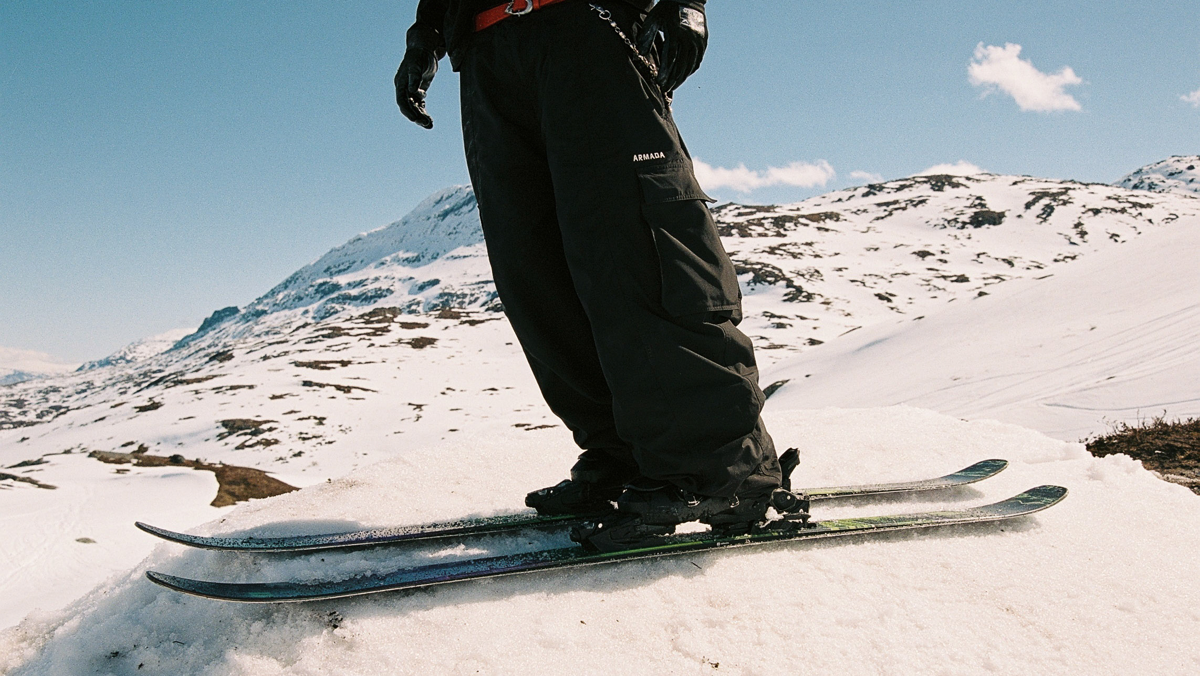 A person wearing black ski pants and gloves stands on skis in a snowy mountain landscape under a blue sky.