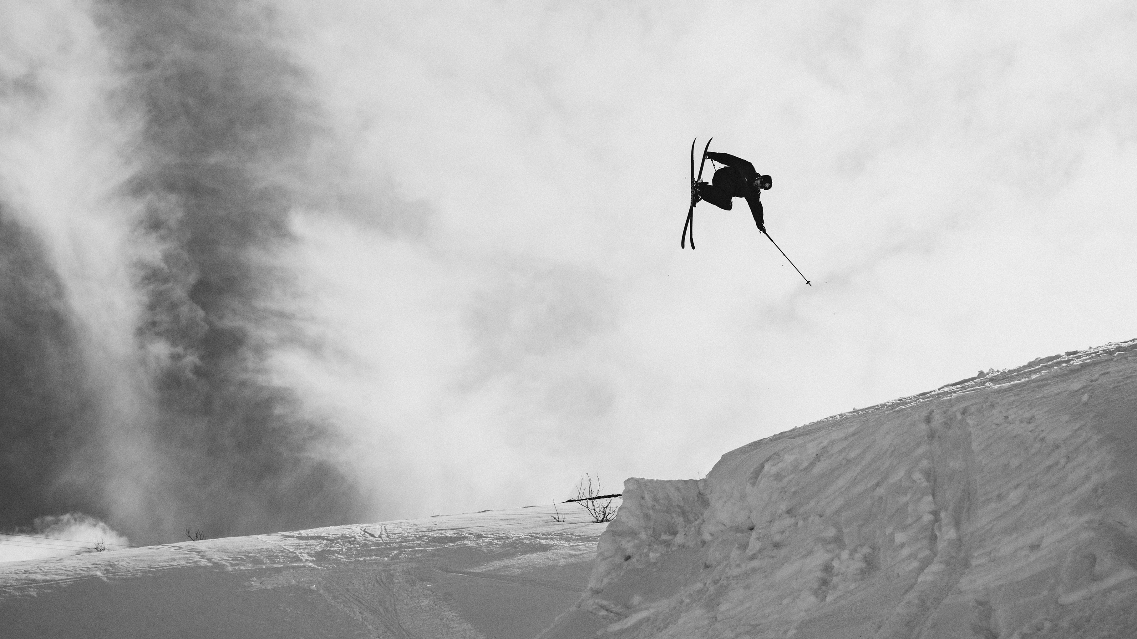 A black and white action shot of a skier mid-air performing a jump over a snowy slope, with a large plume of steam or smoke in the background.