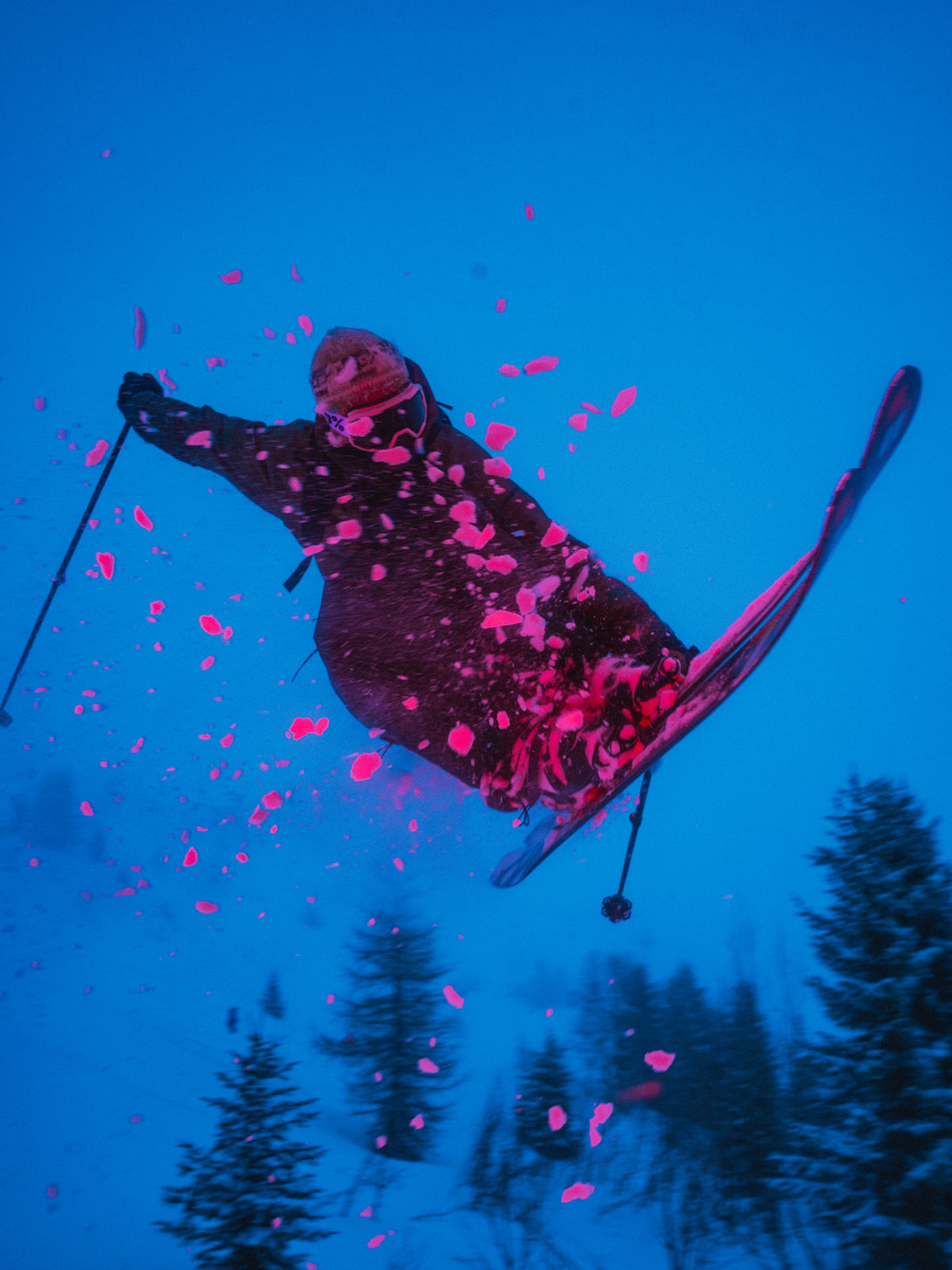 A skier mid-air performing a jump in a snowy mountain setting under a deep blue sky, with bright pink snow petals scattered around them.