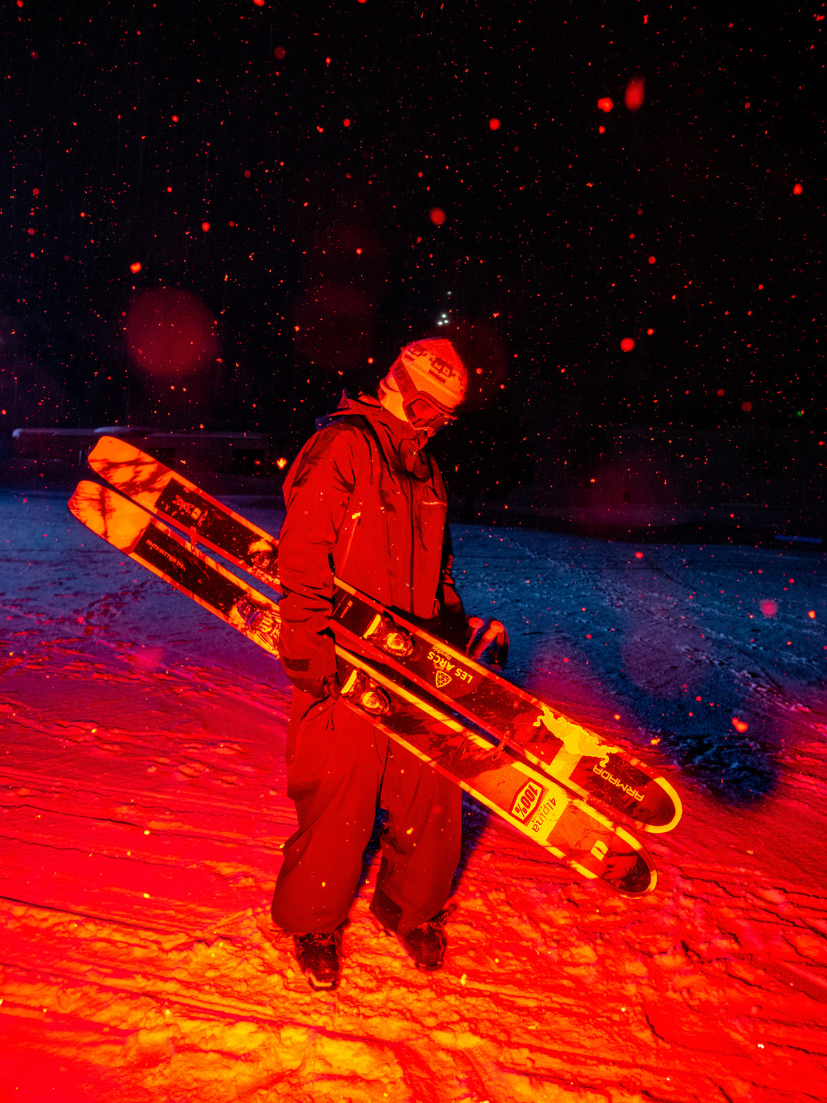 A person in ski gear stands in snow at night carrying a pair of skis under their arm, lit dramatically by red and orange light against a dark sky with visible snowflakes.