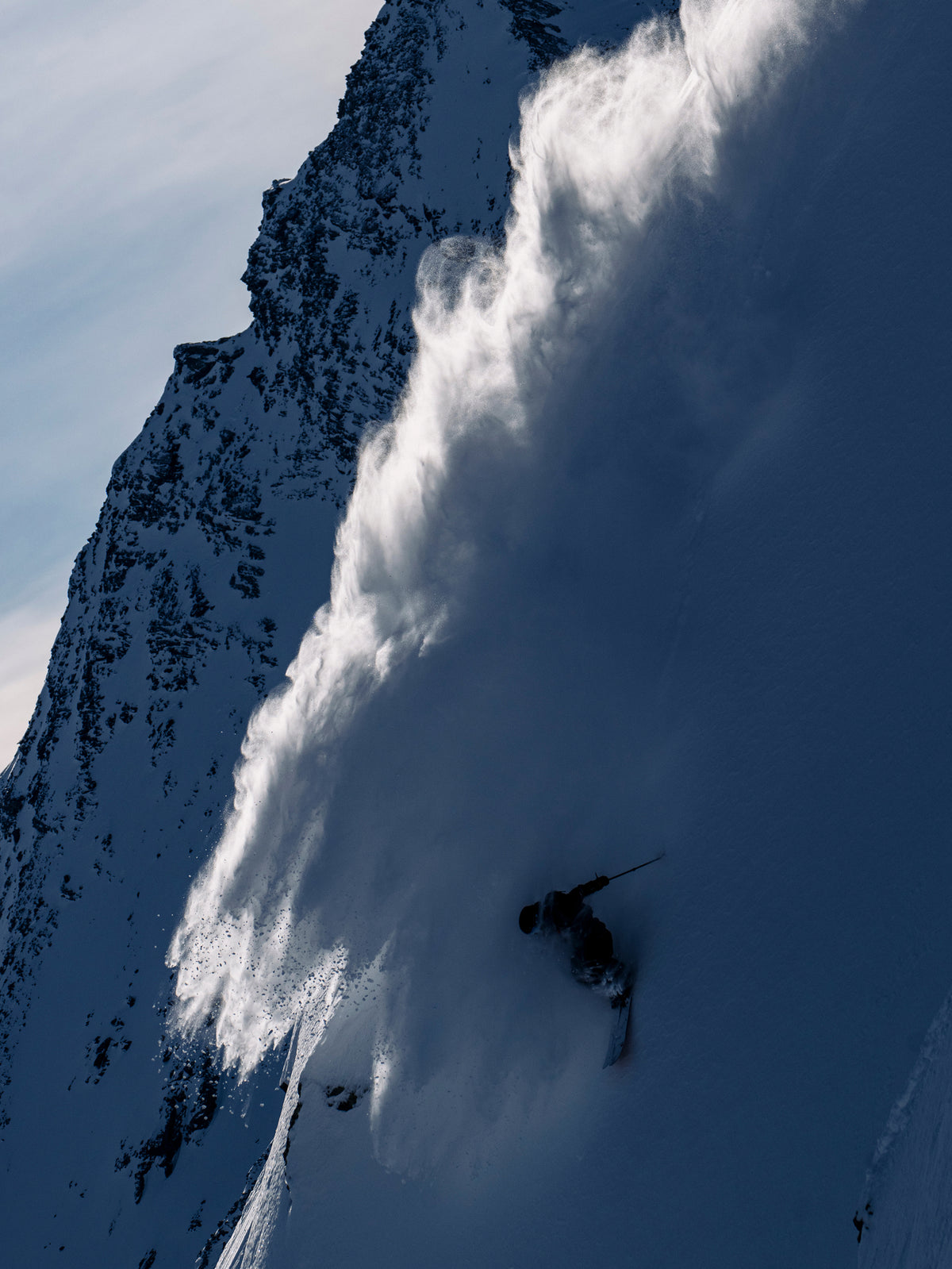 A skier carving through deep powder snow on a steep mountain slope, with a large plume of snow spraying upward and the sky visible in the upper left.