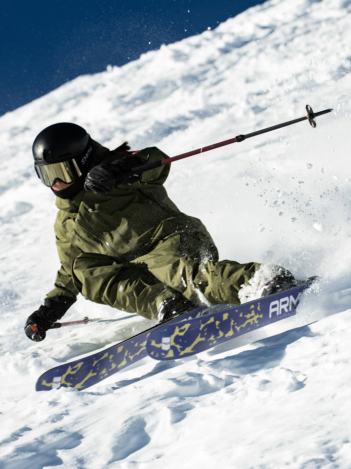 A skier in an olive green snowsuit and black helmet carving through fresh powder on a snowy mountain slope under a clear blue sky. The skier is using ski poles and a blue and yellow patterned ski.