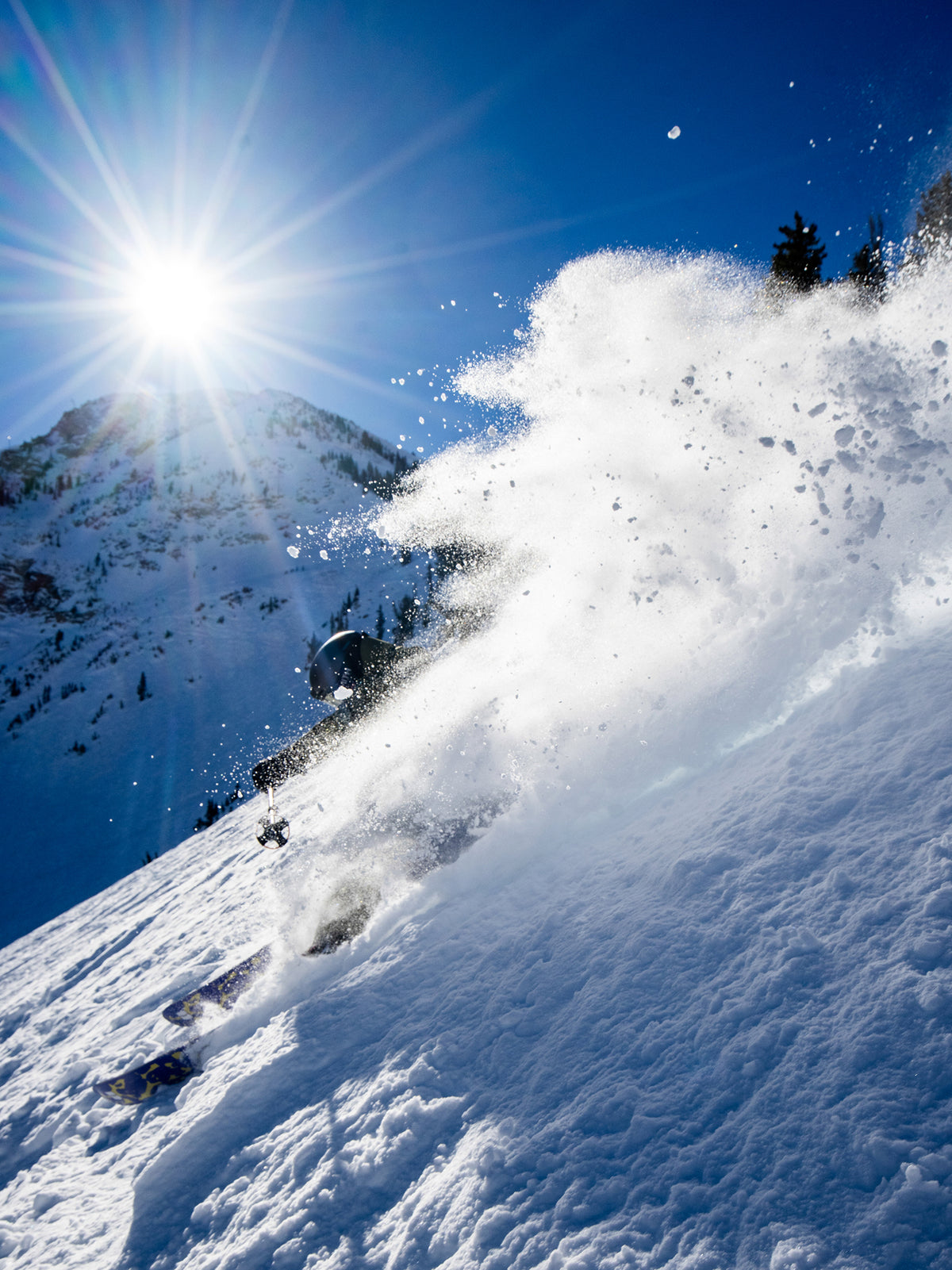 A skier carving through deep powder snow on a sunlit mountain slope, with a large spray of snow in the foreground and a bright sun in a blue sky.