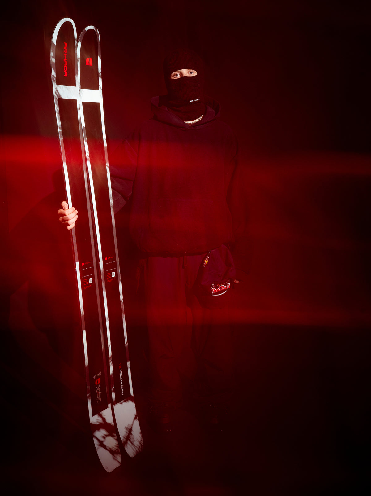A person in a black balaclava and dark outfit holds a pair of white and black skis against a dark red-lit background.
