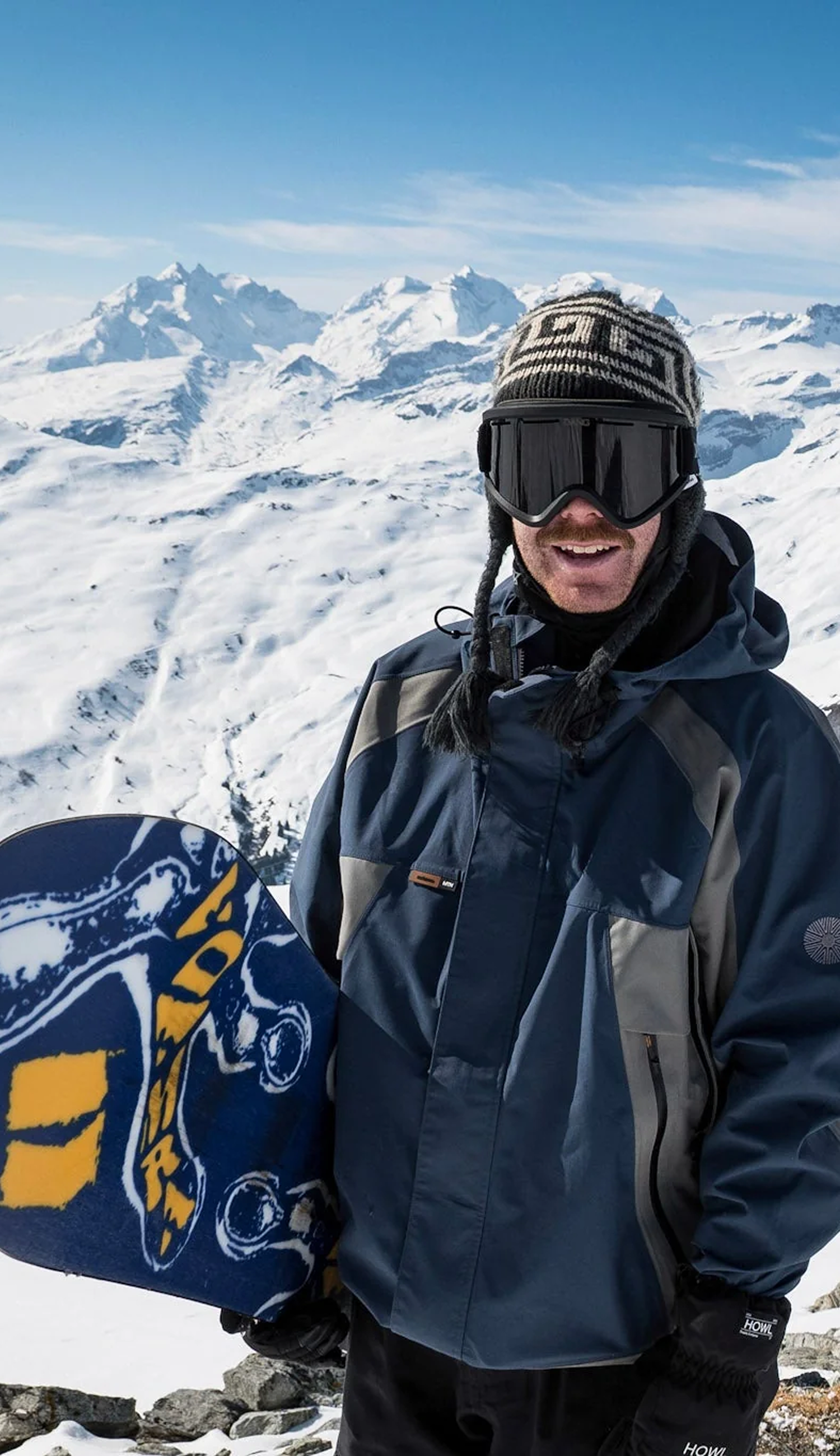 A person wearing a navy and gray ski jacket, black goggles, and a knit beanie stands on a snowy mountain holding a blue and yellow snowboard. The background shows a vast snowy landscape and distant peaks under a blue sky.