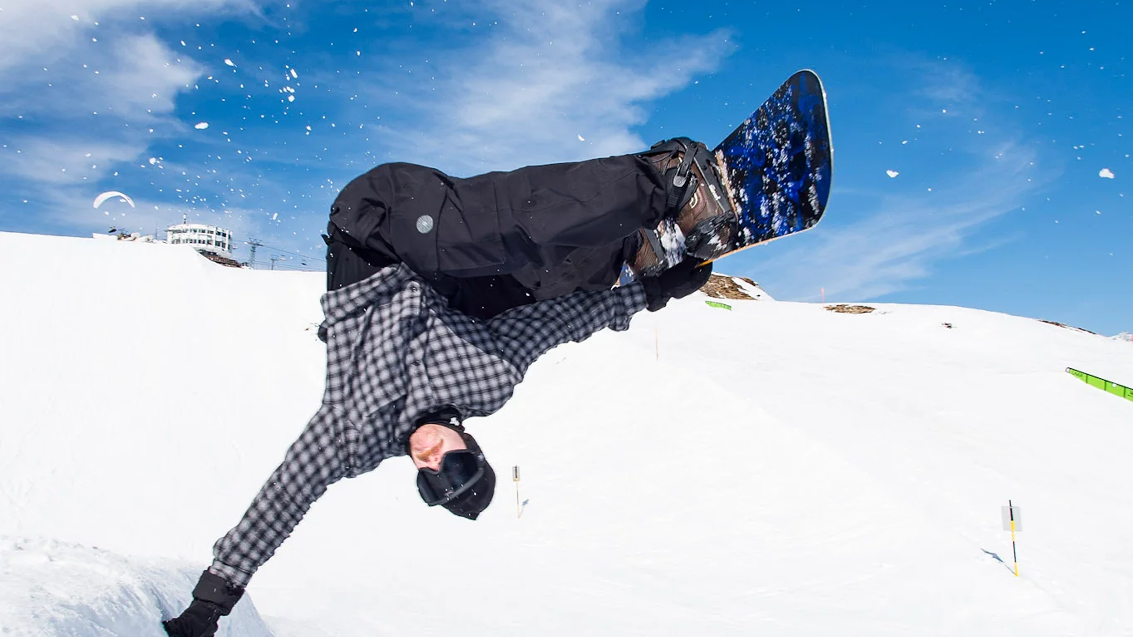 A snowboarder in a black and gray plaid jacket and black pants performs a dynamic flip mid-air on a snowy mountain under a bright blue sky, with a snowboard featuring a blue and black graphic pattern.