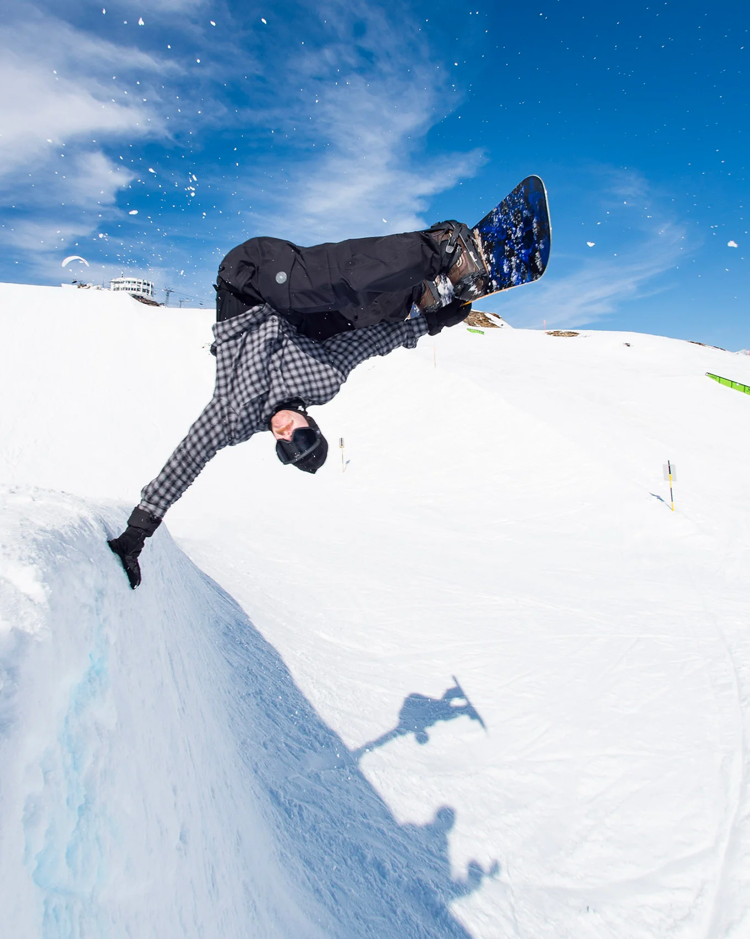 A snowboarder mid-air performing an inversion trick on a snowy mountain slope under a bright blue sky with light clouds.