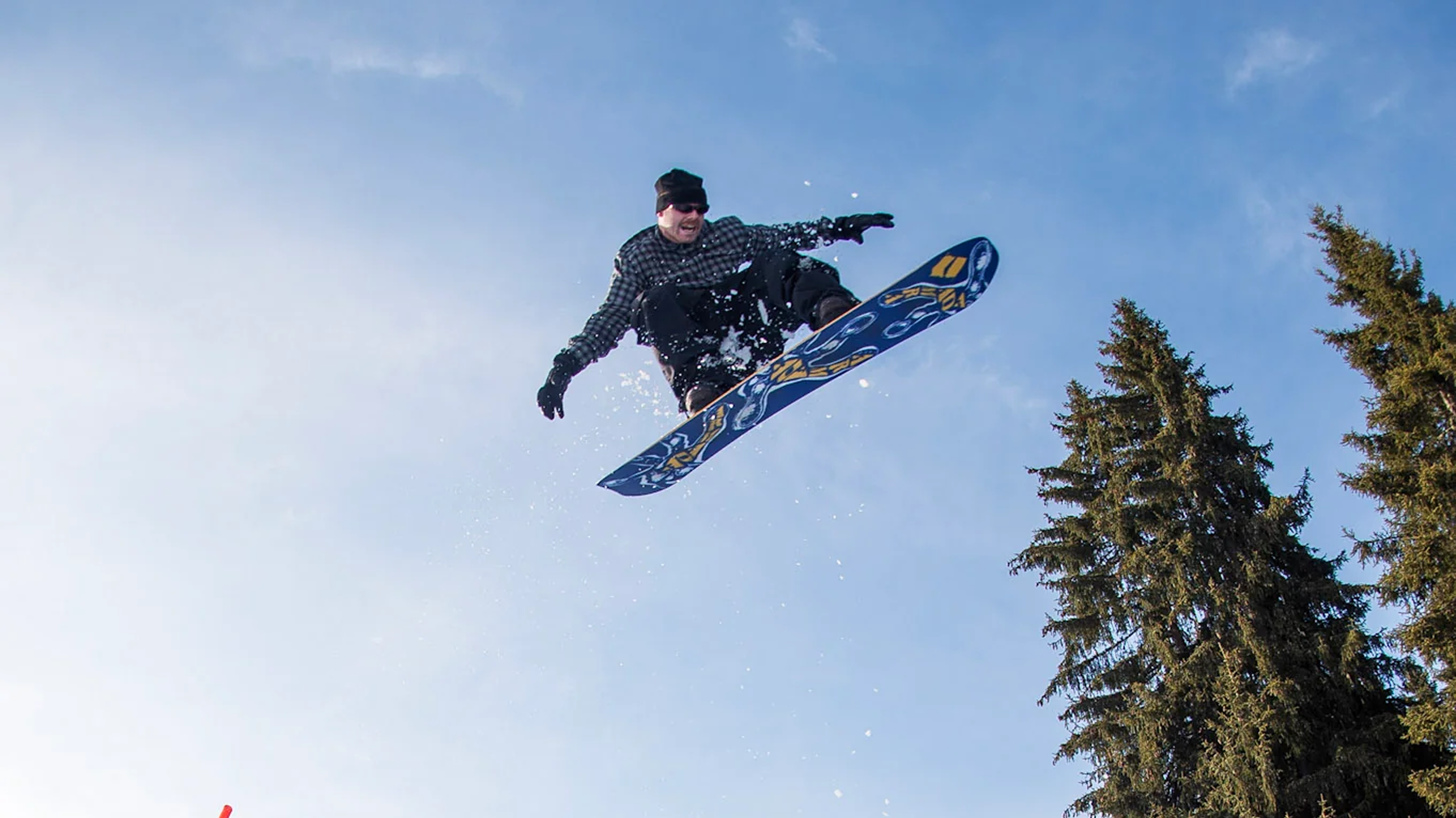 A snowboarder mid-air performing a jump against a blue sky, with evergreen trees on the right side of the frame.