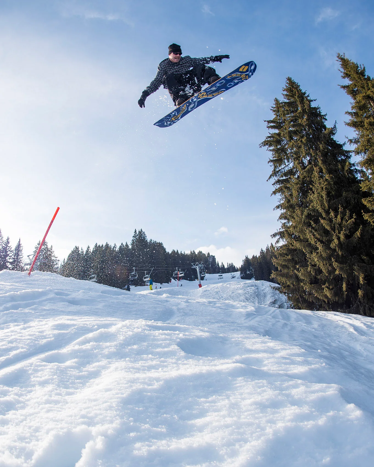 A snowboarder in a black outfit jumps a blue and yellow snowboard over a snowy slope at a ski resort, with trees and a blue sky in the background.