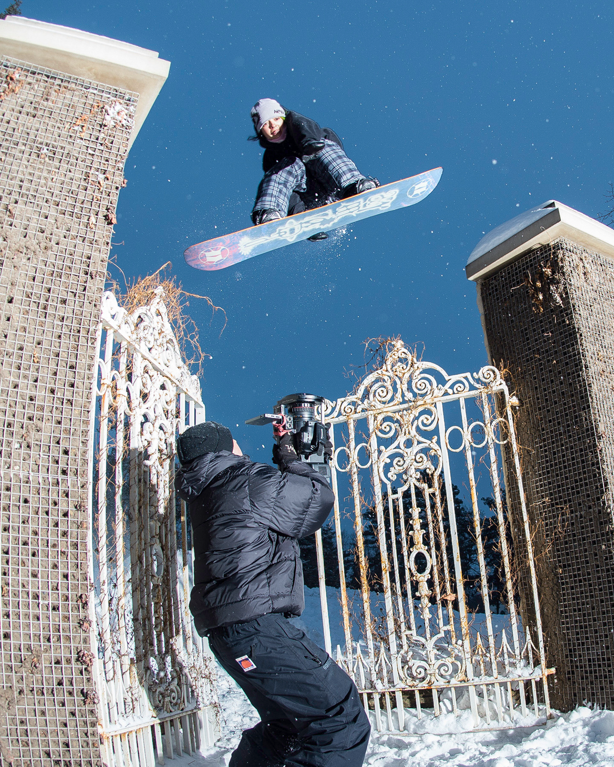A snowboarder mid-air performing a jump in a snowy outdoor setting, with a photographer capturing the moment below between two brick gate pillars under a clear blue sky.