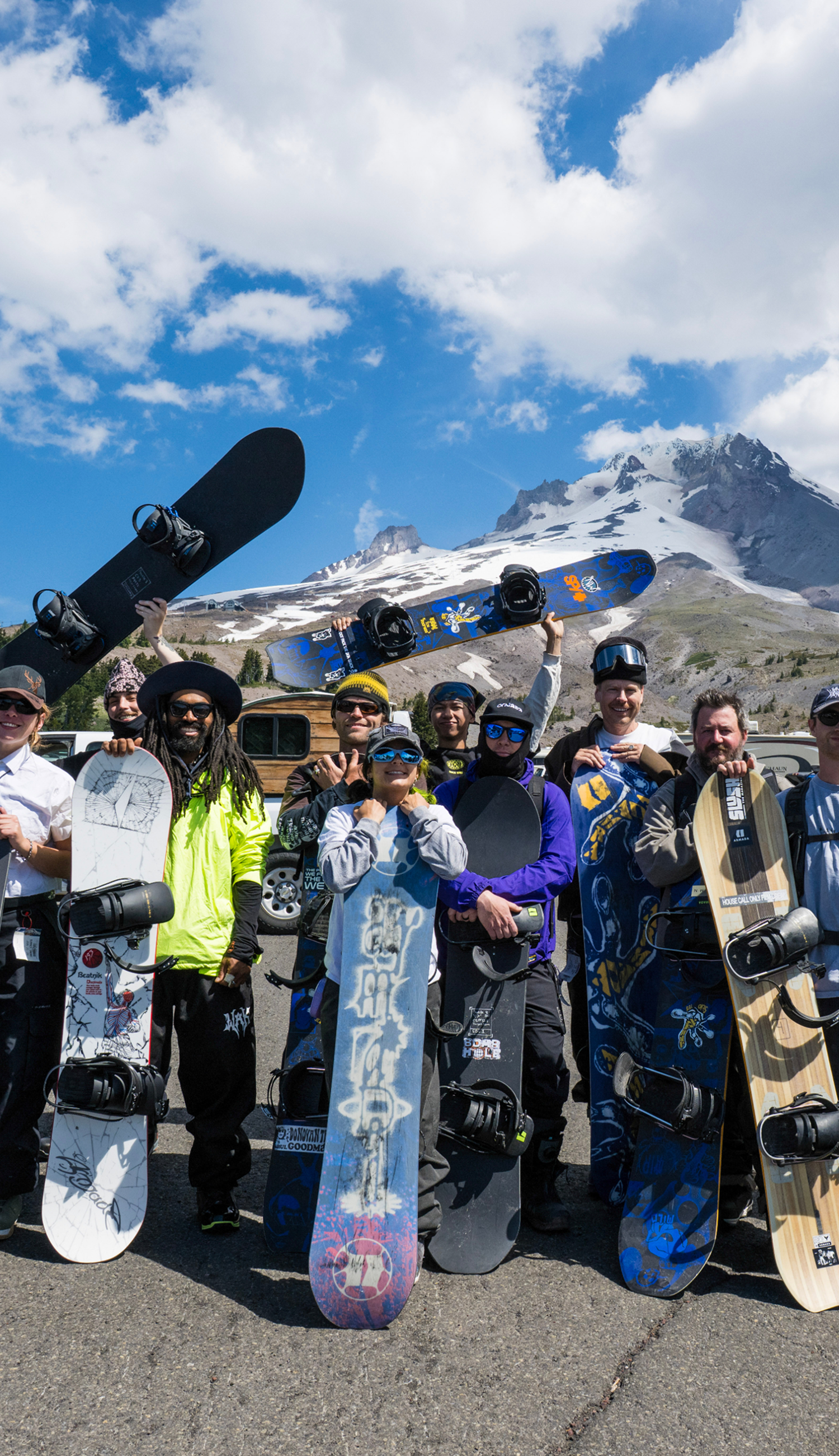 A group of snowboarders posing outdoors in front of a snowy mountain under a blue sky with clouds, each holding up a snowboard. The scene is casual and recreational, with people wearing winter sports gear and helmets.