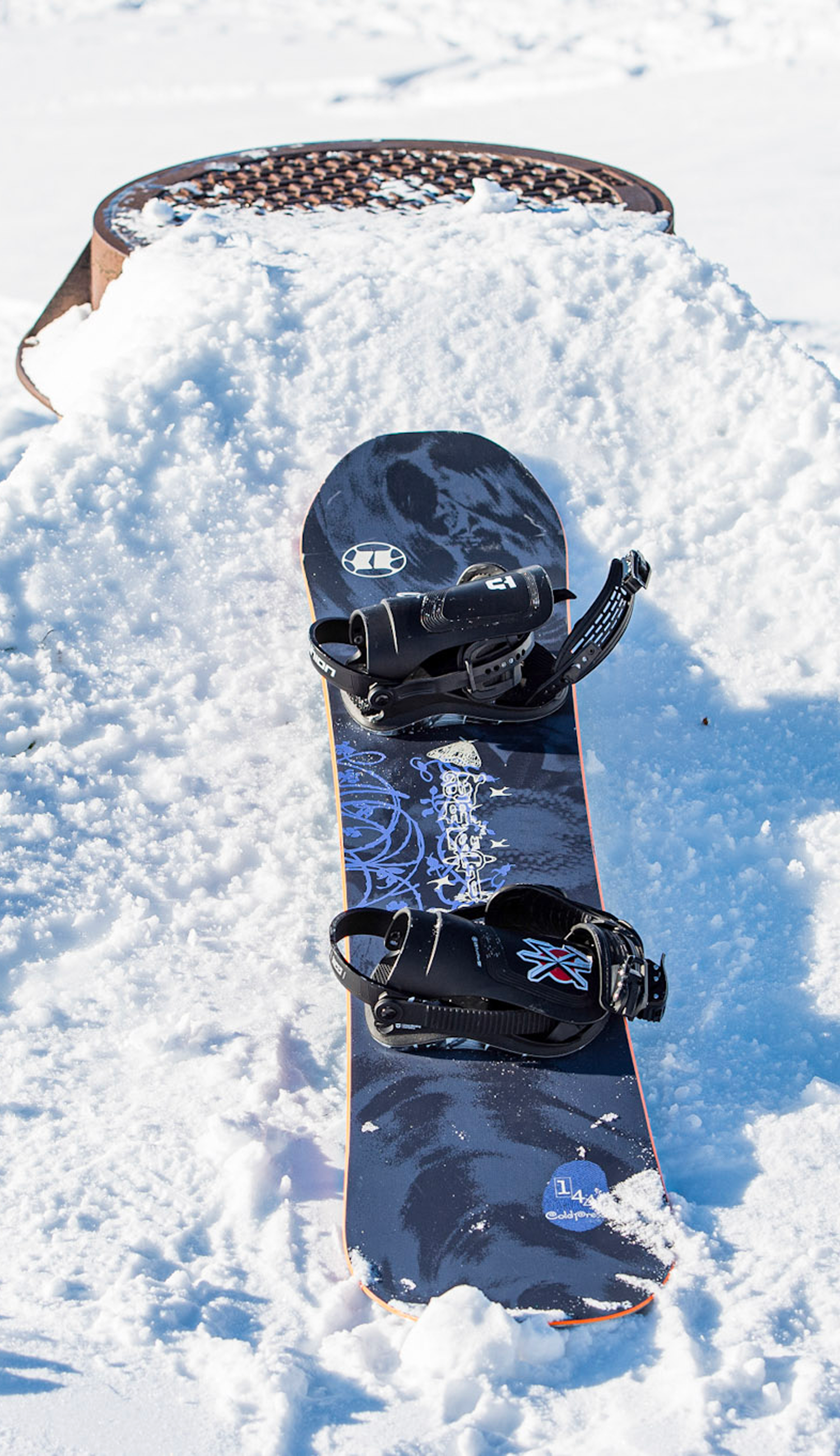 A black snowboard with blue graphics and black bindings sits upright in deep snow, with a metal snowboard rack partially visible behind it.