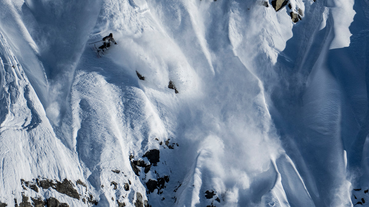 Snow-covered mountain slopes with dramatic wind-blown powder and dark rocky outcrops in a high-altitude alpine setting.