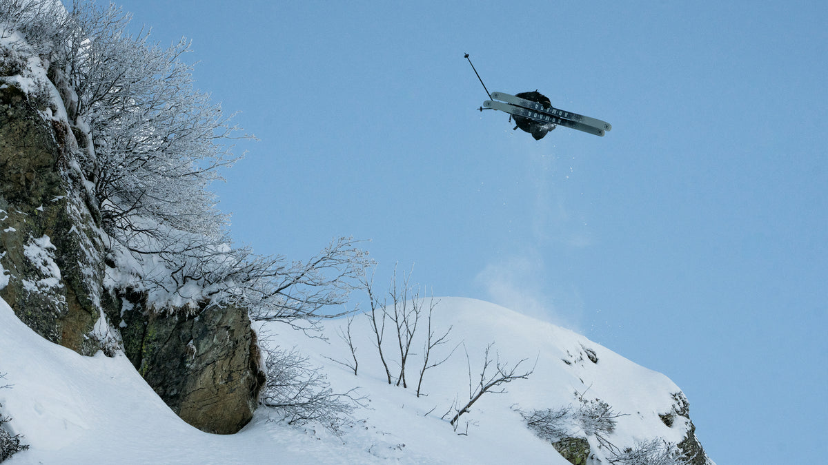 A skier in mid-air performing a jump over a snowy mountain slope with bare trees and a rocky cliff under a clear blue sky.